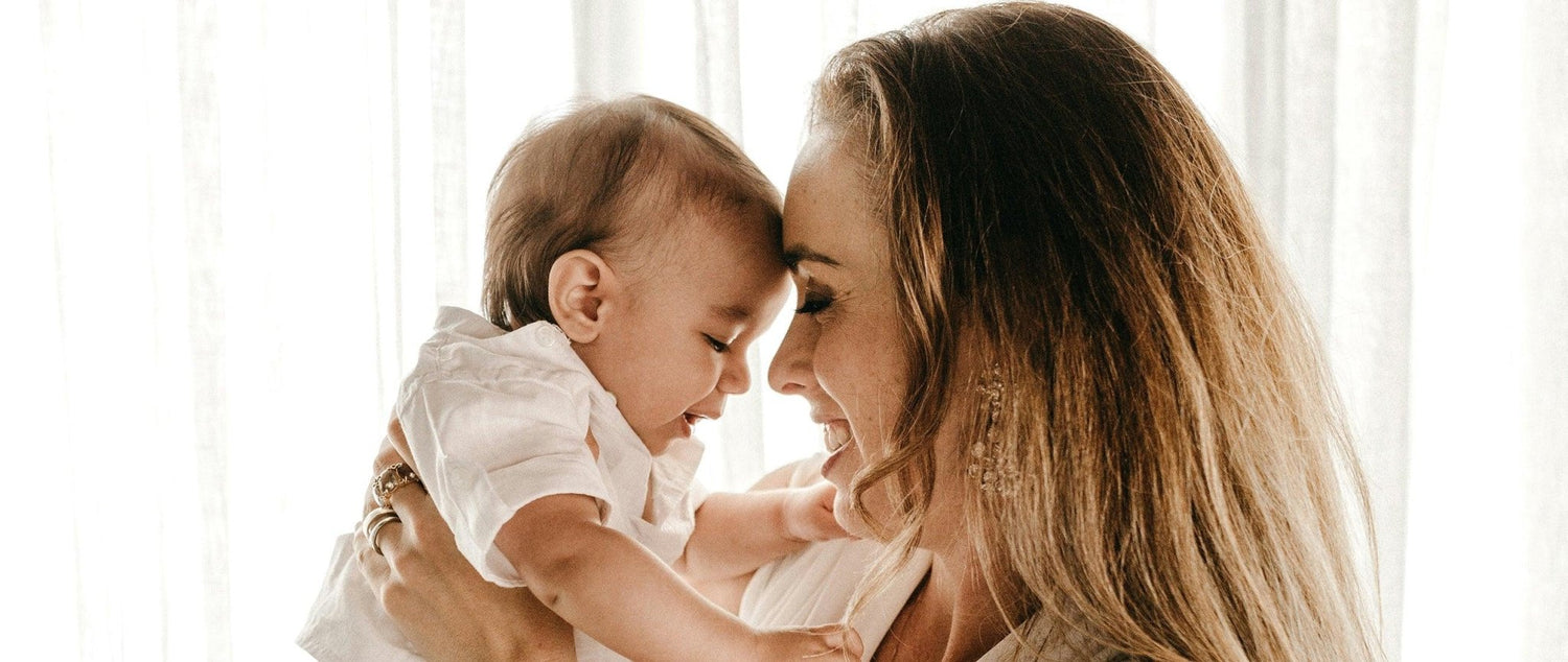 Maman souriante portant un bébé, complicité et tendresse, lumière naturelle en fond blanc